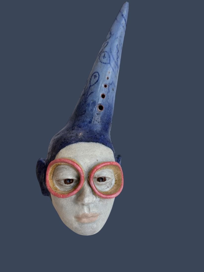 Daydreamer, a ceramic wall mask by Ireri Krueger of a pale face in oversized pink-rimmed glasses beneath a tall, doodle-covered blue hat
