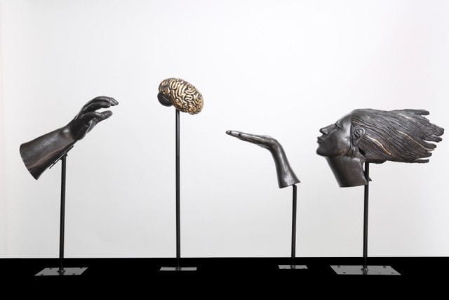 Fragments, a ceramic installation by Ireri Krueger of a hand, a brain, a blown kiss, and a windblown head displayed on slender black stands