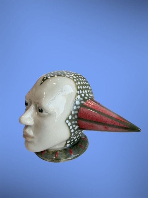 Migration, a ceramic sculpture by Ireri Krueger of a serene profile head fitted with a long red-and-green striped bird beak